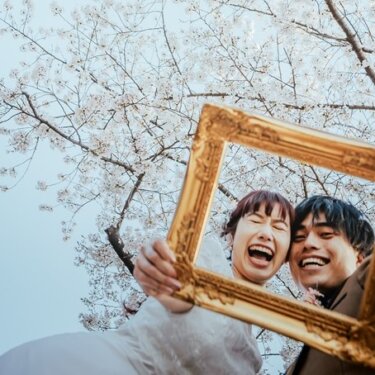 満開の桜の下で金色の額縁を掲げ、顔を寄せて笑い合う新郎新婦。遊び心のある額縁ポーズで撮る桜の前撮り