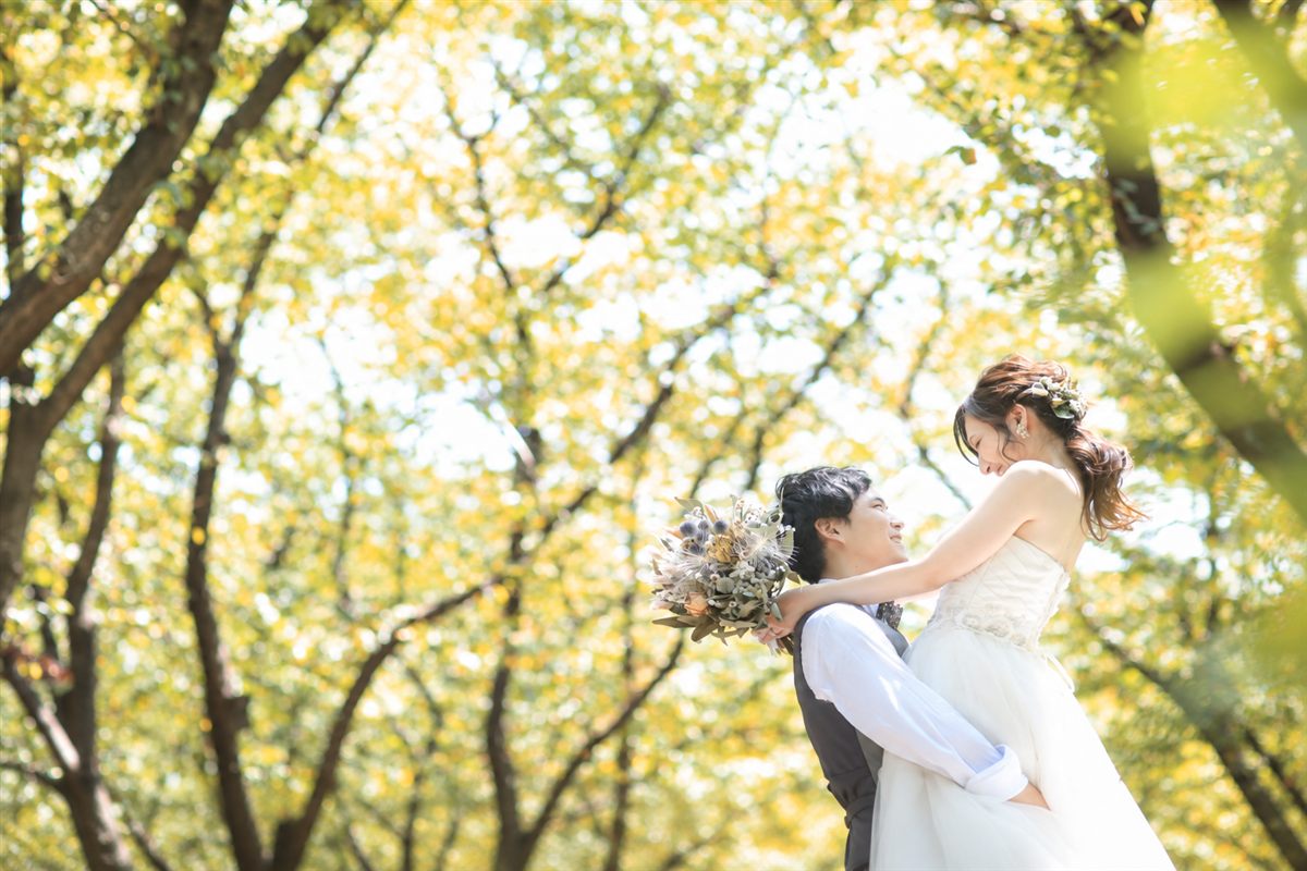 黄色く色づいた木々の下でウェディングドレスの花嫁を抱き上げるベスト姿の新郎