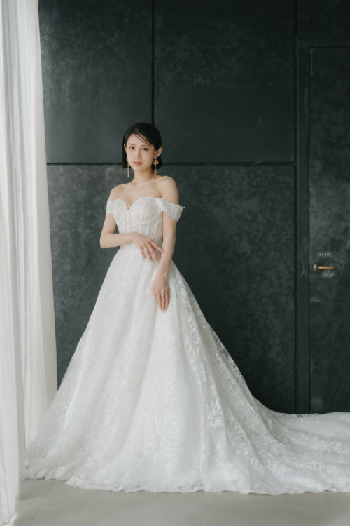 ダークな壁を背に腕を軽く組む花嫁。花柄レースが広がるプリンセスラインのウェディングドレスDC02で撮る洋装前撮り
