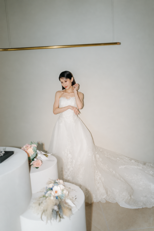 花を飾った円柱の台の横で、耳元に手を添えてポーズをとるAラインのウェディングドレスの花嫁。DC08のスタジオフォト
