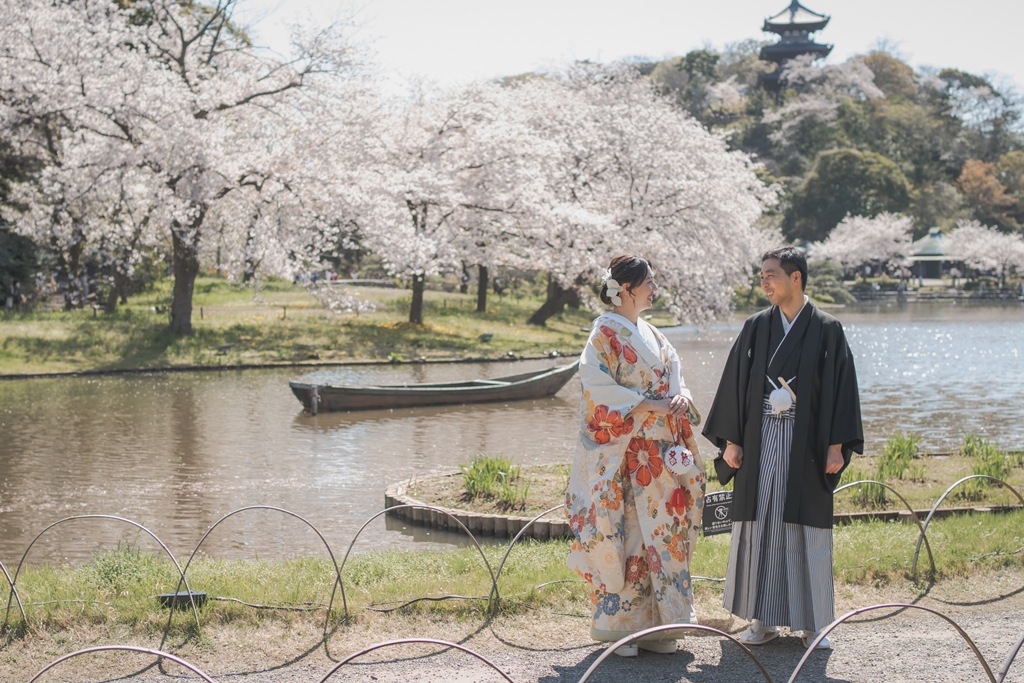 満開の桜と池に浮かぶ小舟、遠くの三重塔を望む三渓園で、向かい合う色打掛の花嫁と羽織袴の新郎を引きで写した和装前撮り