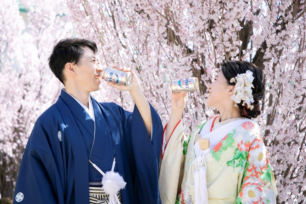 満開の桜の下で缶ジュースを手に乾杯する紺の紋付袴の新郎と色打掛の花嫁。遊び心のある和装前撮り