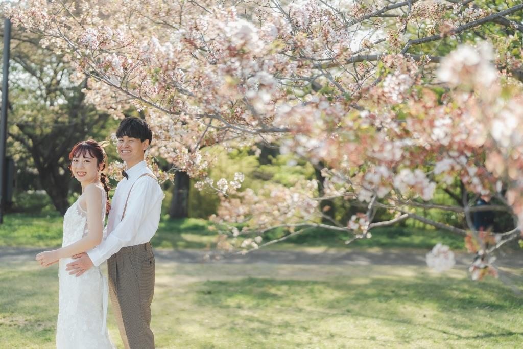 八重桜の咲く公園で、花嫁を後ろから抱きしめて笑う白いシャツの新郎。桜で撮る洋装前撮りのポーズ