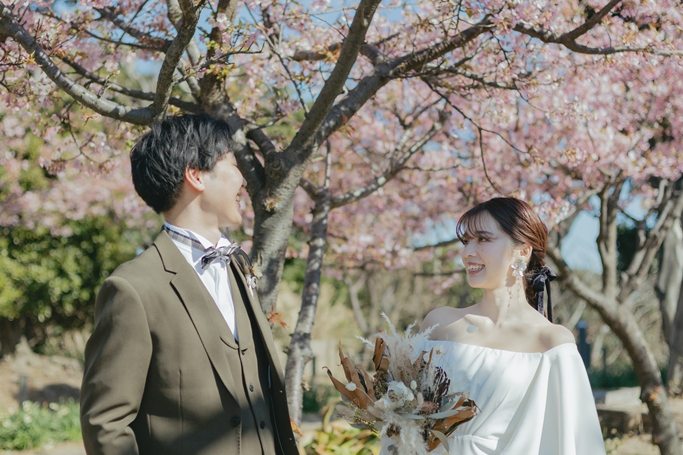 満開の桜を背に見つめ合う、ブラウンのスーツの新郎とウェディングドレスの花嫁。桜で撮る洋装前撮り