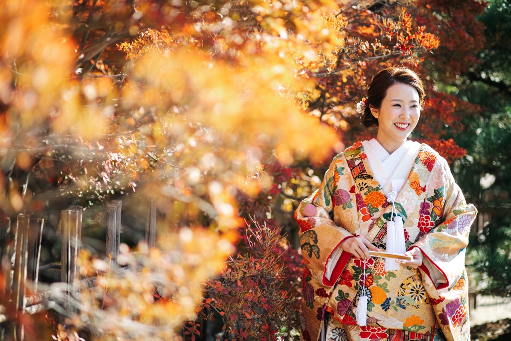 色づいた紅葉の木々の中でほほえむウェディングドレスの花嫁。秋の紅葉前撮りで人気の洋装ロケーションフォト