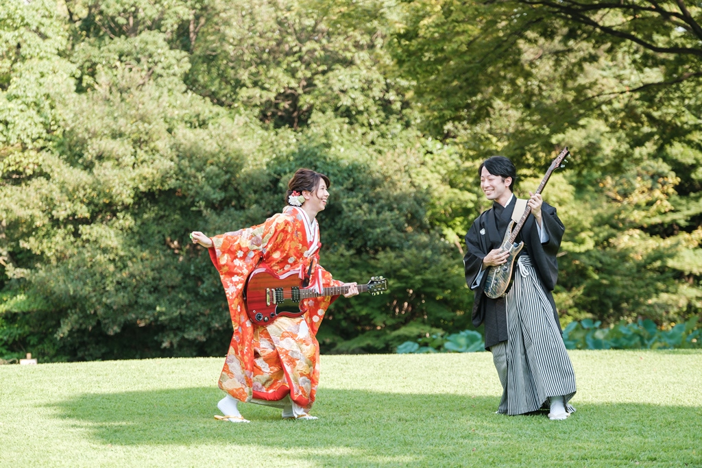 緑の芝生でエレキギターを手に笑い合う色打掛の新婦と紋付袴の新郎。9月の緑が残る季節に撮る個性的な和装前撮り