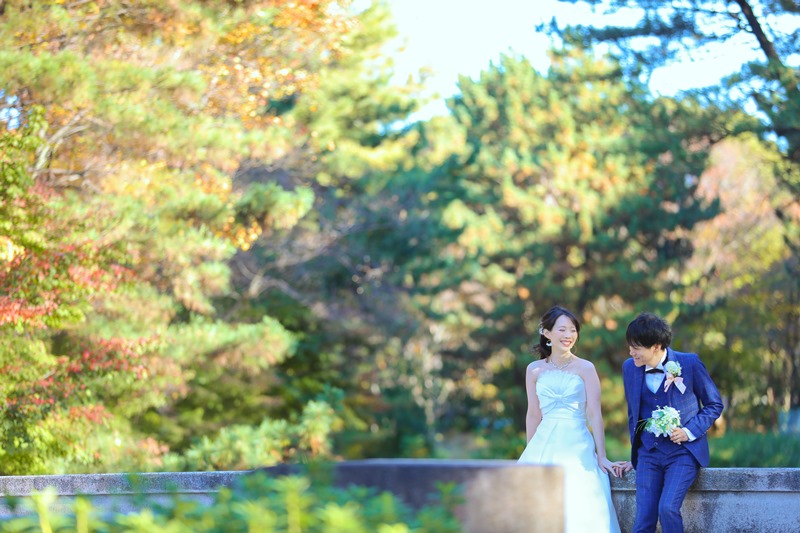 色づき始めた木々を背景に石の欄干に座るウェディングドレスの新婦と青いタキシードの新郎。10月の秋色の洋装前撮り