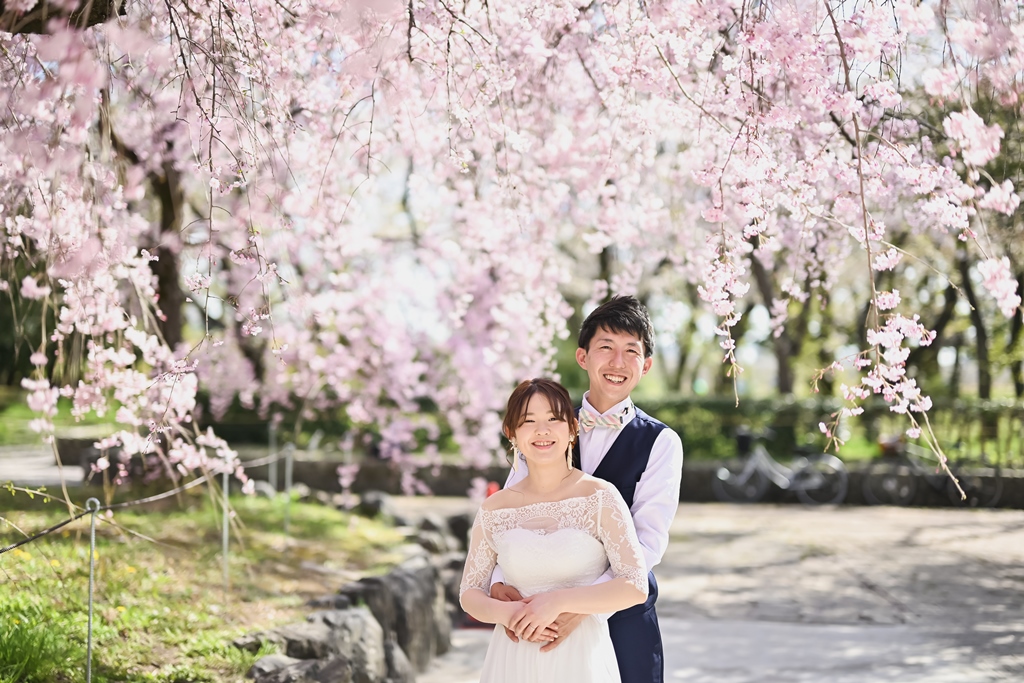 桜の木の下で笑顔を見せるウェディングドレスの新婦とタキシードの新郎。春のフォトウェディングらしい明るい結婚写真