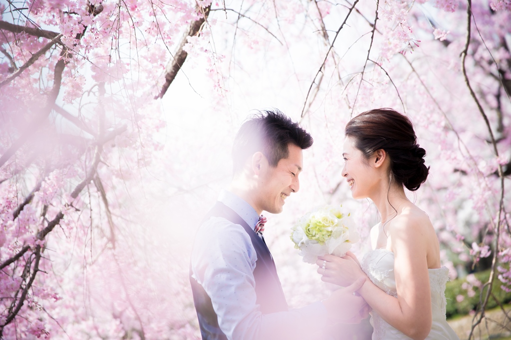満開の桜を背景に寄り添うウェディングドレスの新婦とタキシードの新郎。春の桜前撮りで人気の洋装ロケーションフォト