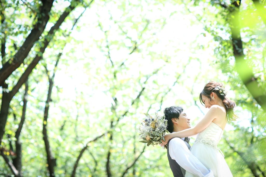 新緑の公園でウェディングドレスの新婦を抱き上げるタキシードの新郎。春の洋装前撮りで人気のお姫様抱っこ