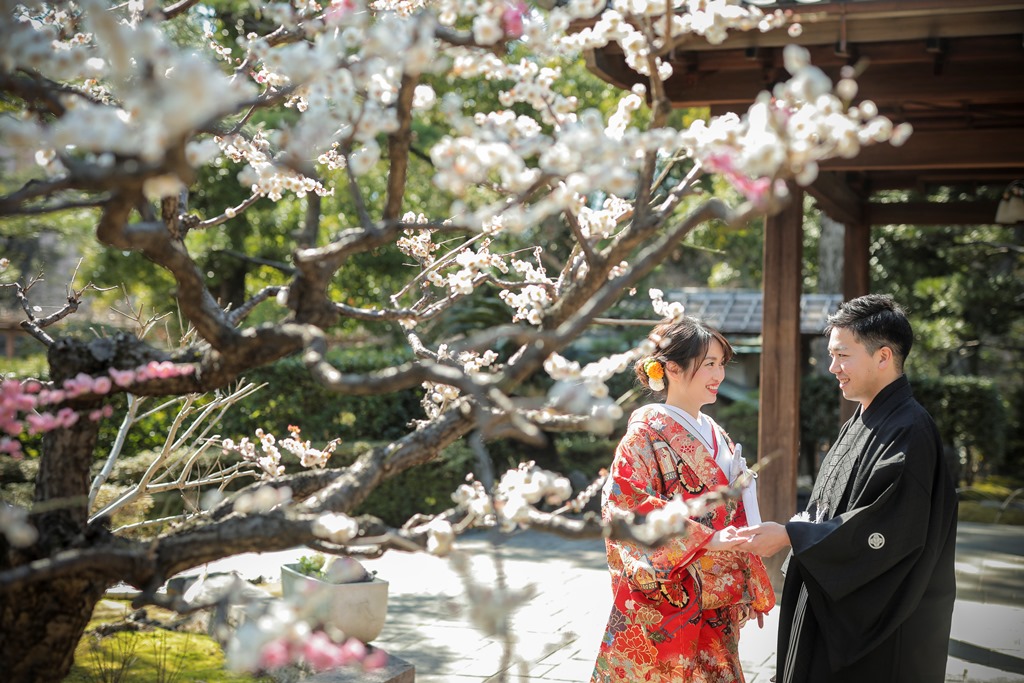 白梅の咲く庭園で向かい合う赤い花柄の振袖姿の花嫁と黒紋付袴の新郎。3月の梅と撮る和装前撮り