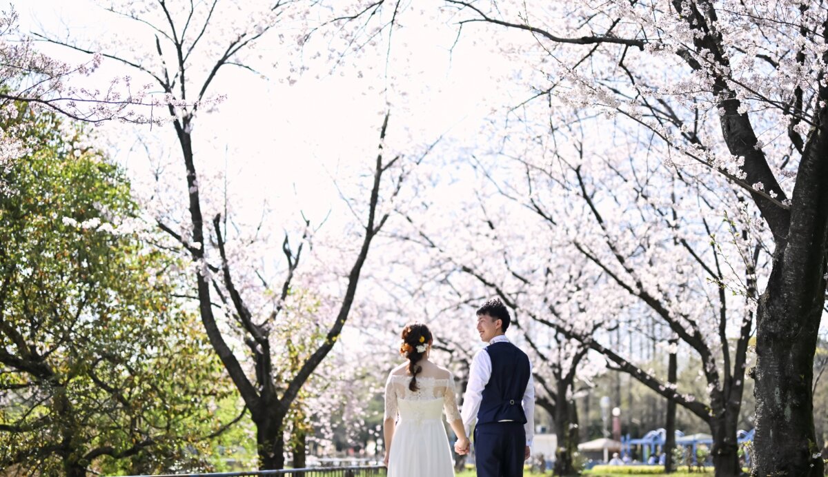 満開の桜並木を手をつないで歩くレースのウェディングドレスの新婦とベスト姿の新郎の後ろ姿。桜前撮りで人気の自然なポーズ