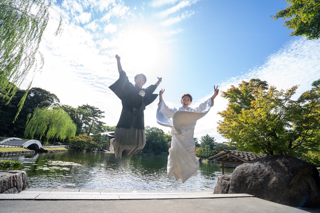 日本庭園の池のほとりで青空に向かってジャンプする白無垢の新婦と紋付袴の新郎。夏の和装前撮りで人気の元気なポーズ