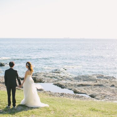 海を望む岩場の芝生で手をつなぐウェディングドレスの新婦とタキシードの新郎の後ろ姿。夏の海で撮る洋装前撮り