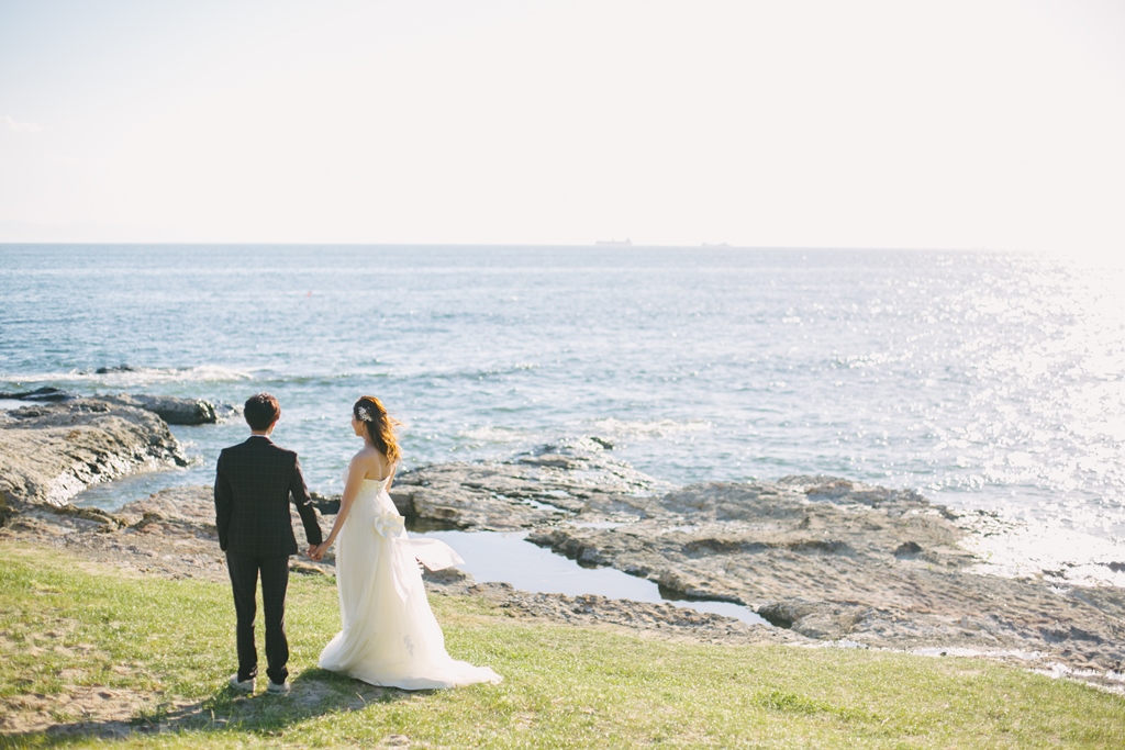 海を望む岩場の芝生で手をつなぐウェディングドレスの新婦とタキシードの新郎の後ろ姿。夏の海で撮る洋装前撮り