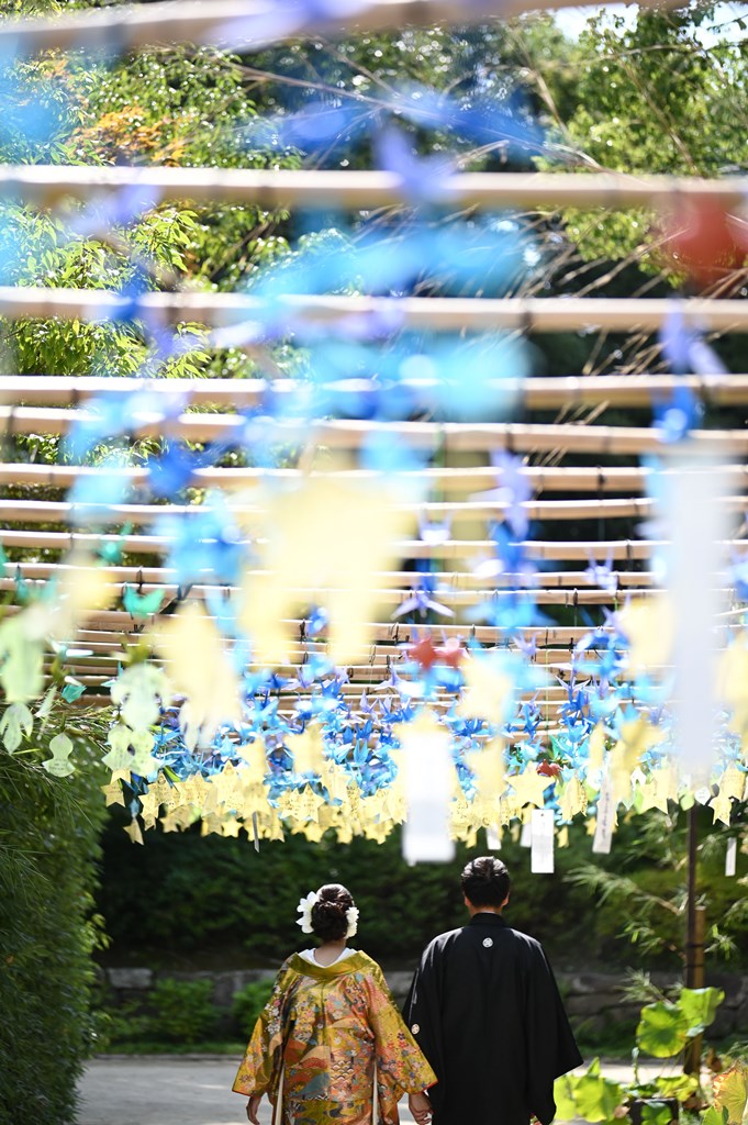 七夕の吹き流し飾りの下を歩く花柄の着物姿の花嫁と黒紋付袴の新郎。7月の七夕に撮る和装前撮り