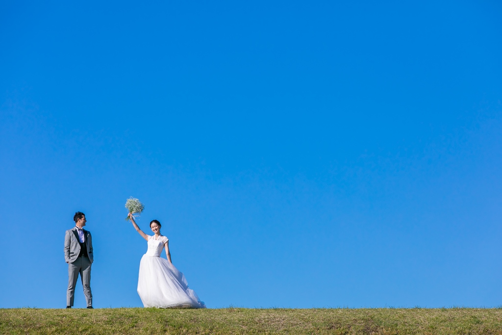 青空の下の土手でブーケを掲げるウェディングドレスの新婦とグレーのタキシードの新郎。夏の明るい光で撮る洋装ロケーションフォト