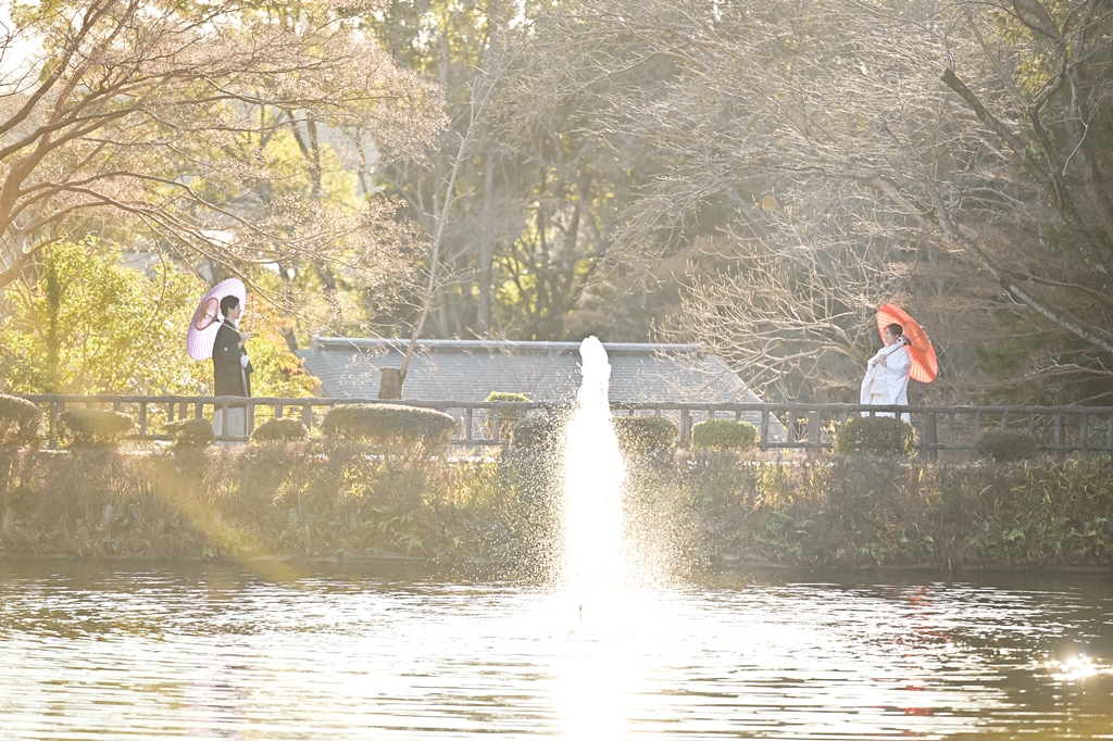 噴水のある池をはさんで和傘を手に向かい合う白無垢の新婦と紋付袴の新郎。冬の澄んだ光に包まれる和装前撮り