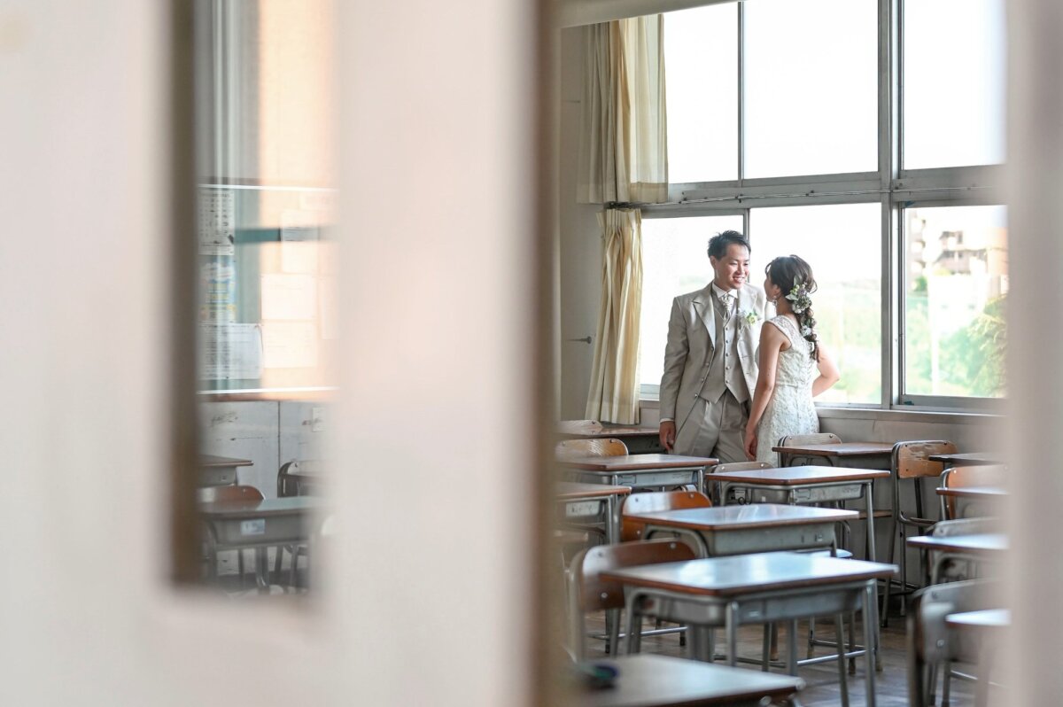 教室の窓辺で見つめ合うグレーのタキシードの新郎とレースのウェディングドレスの新婦。思い出の学校で撮るフォトウェディング