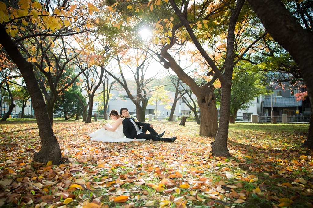 落ち葉の絨毯の上に並んで座るウェディングドレスの新婦とタキシードの新郎。紅葉が残る12月の洋装前撮り